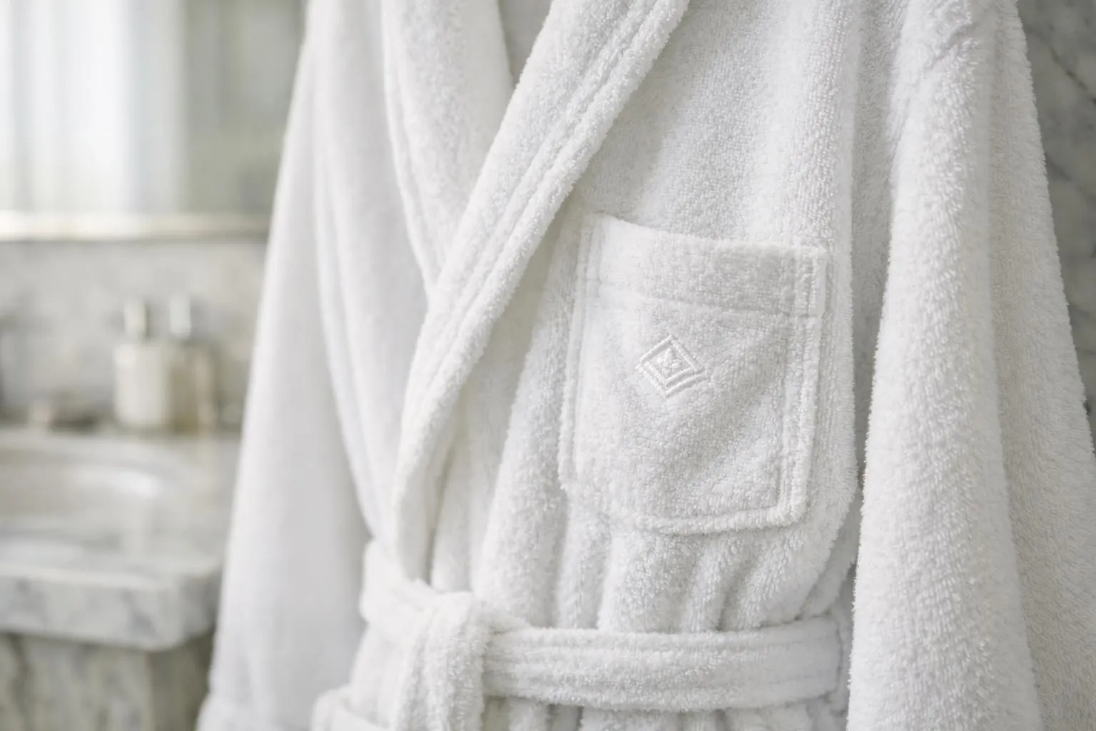 Close-up photograph of an elegant white hotel bathrobe with a discreet embroidered logo on the chest pocket, hanging in a luxurious bathroom with soft natural lighting and marble surfaces visible in the background, showing fine textile detail and premium quality stitching