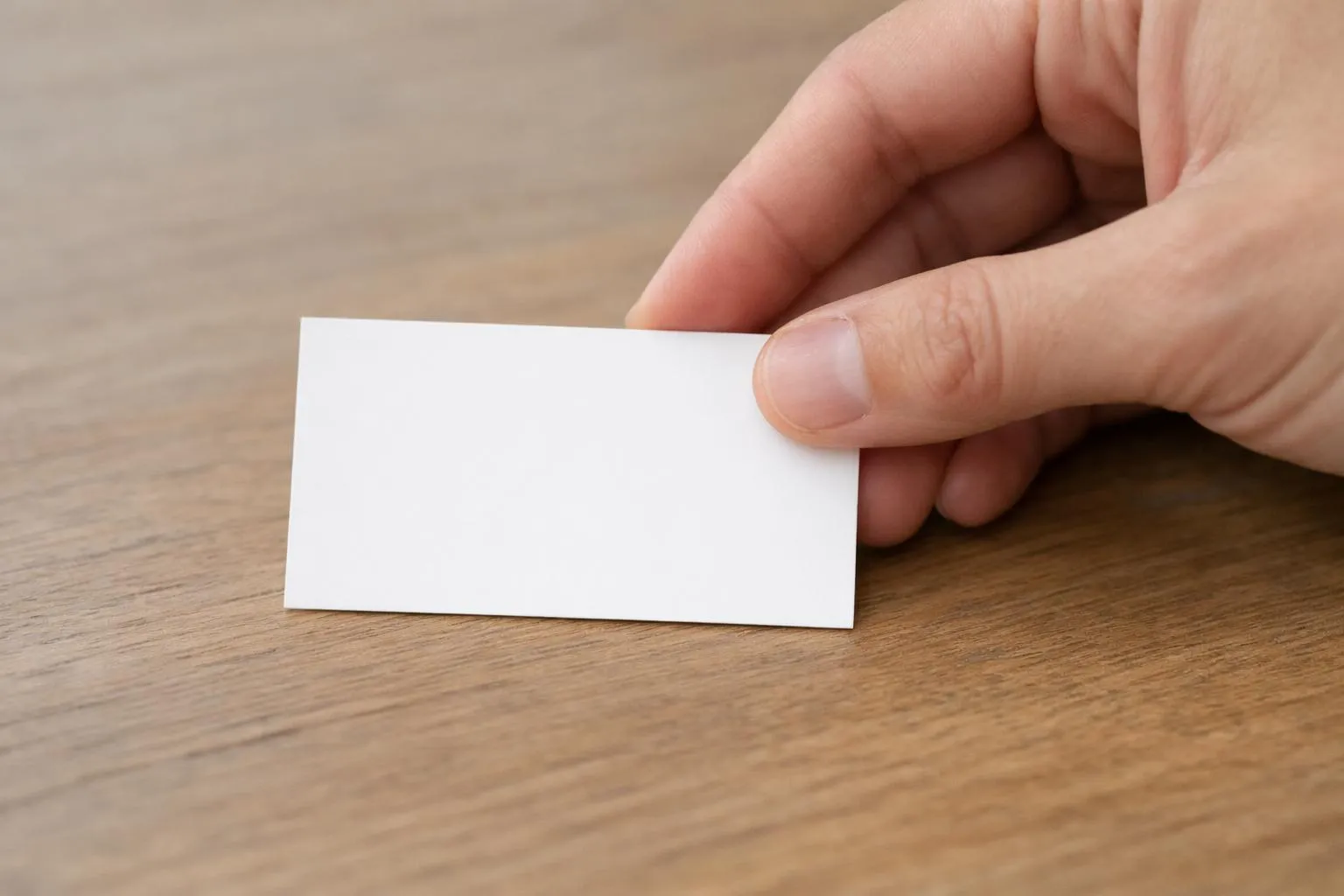 Gros plan d'une main d'homme d'affaires posant une carte de visite blanche minimaliste sur une table en bois clair, la carte présentant uniquement le nom et les coordonnées imprimés en police Helvetica noire, éclairage naturel doux, composition épurée professionnelle, sans aucun texte visible ni logo