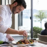 Chef préparant un plat d'agneau grillé dans un restaurant à Lausanne, lumière naturelle et ambiance chaleureuse.