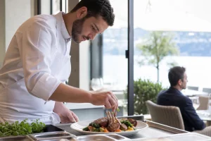 Chef préparant un plat d'agneau grillé dans un restaurant à Lausanne, lumière naturelle et ambiance chaleureuse.