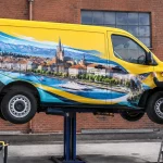 Camion utilitaire original avec covering créatif suspendu devant mur d'atelier à Lausanne, lumière naturelle, conducteur ajus